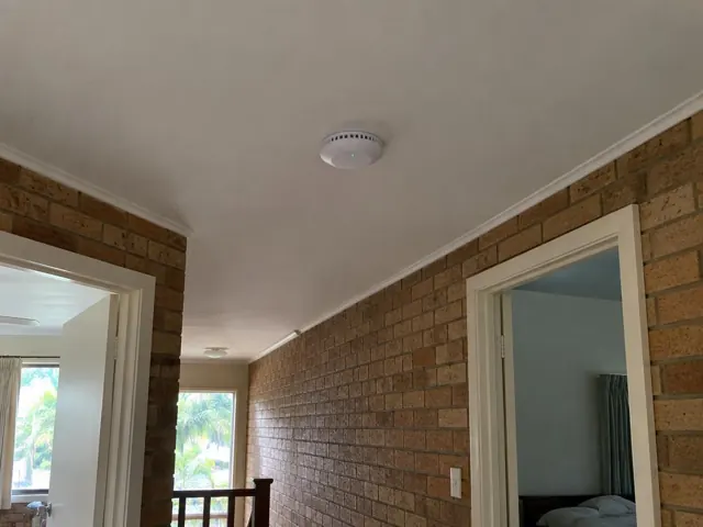 Ceiling smoke alarm in a brick-lined hallway with doorways and white trim in Currumbin Waters